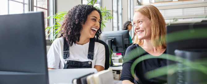 Zwei Frauen sitzen an einem PC und lächeln sich an.