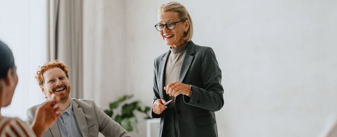 Eine Frau steht im Blazer vor einer Gruppe Mitarbeitenden