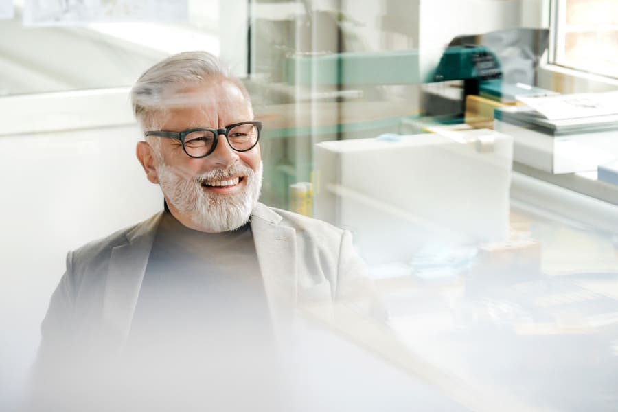 Ein Mann mit grauem Haar und Brille wird durch ein Fenster hindurch fotografiert und lächelt.