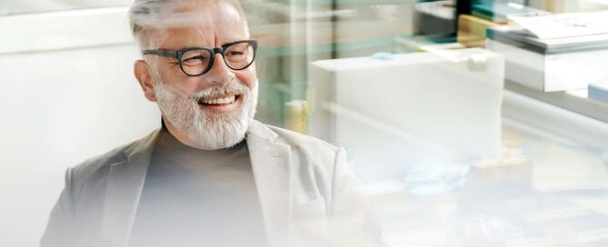 Ein Mann mit grauem Haar und Brille wird durch ein Fenster hindurch fotografiert und lächelt.