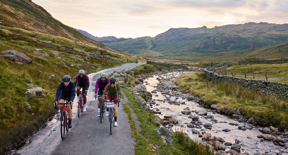 Headerbild einer Gruppe beim Road cycling entlang einer Straße zwischen einem Berg und einem Fluss.